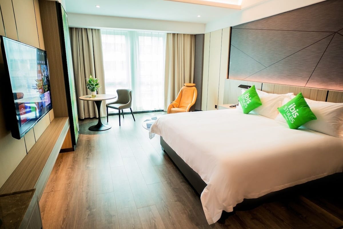 Ein Hotelzimmer mit einem großen Bett, einem an der Wand befestigten Fernseher, einem kleinen Tisch mit Stuhl und einem Fenster mit Vorhängen.
