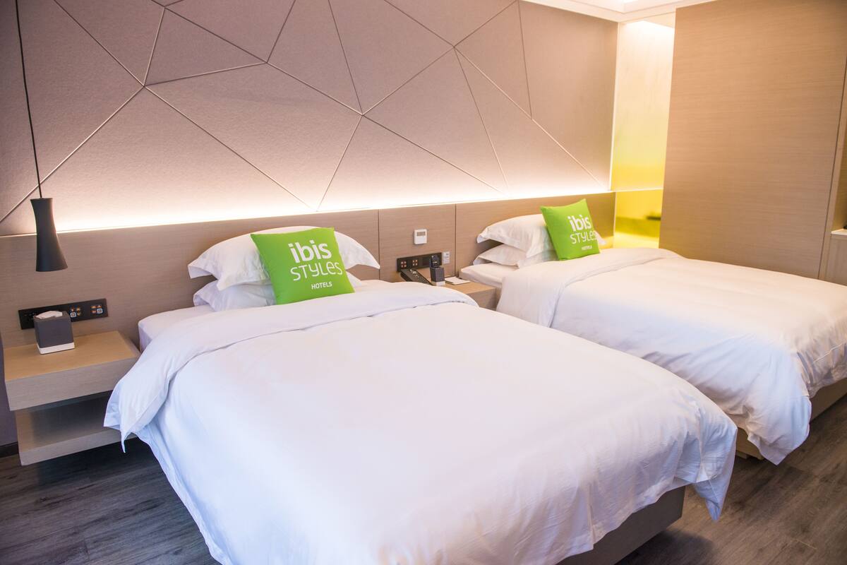 Habitación de hotel con dos camas, una mesita de noche con lámpara y una pared con diseños geométricos.
