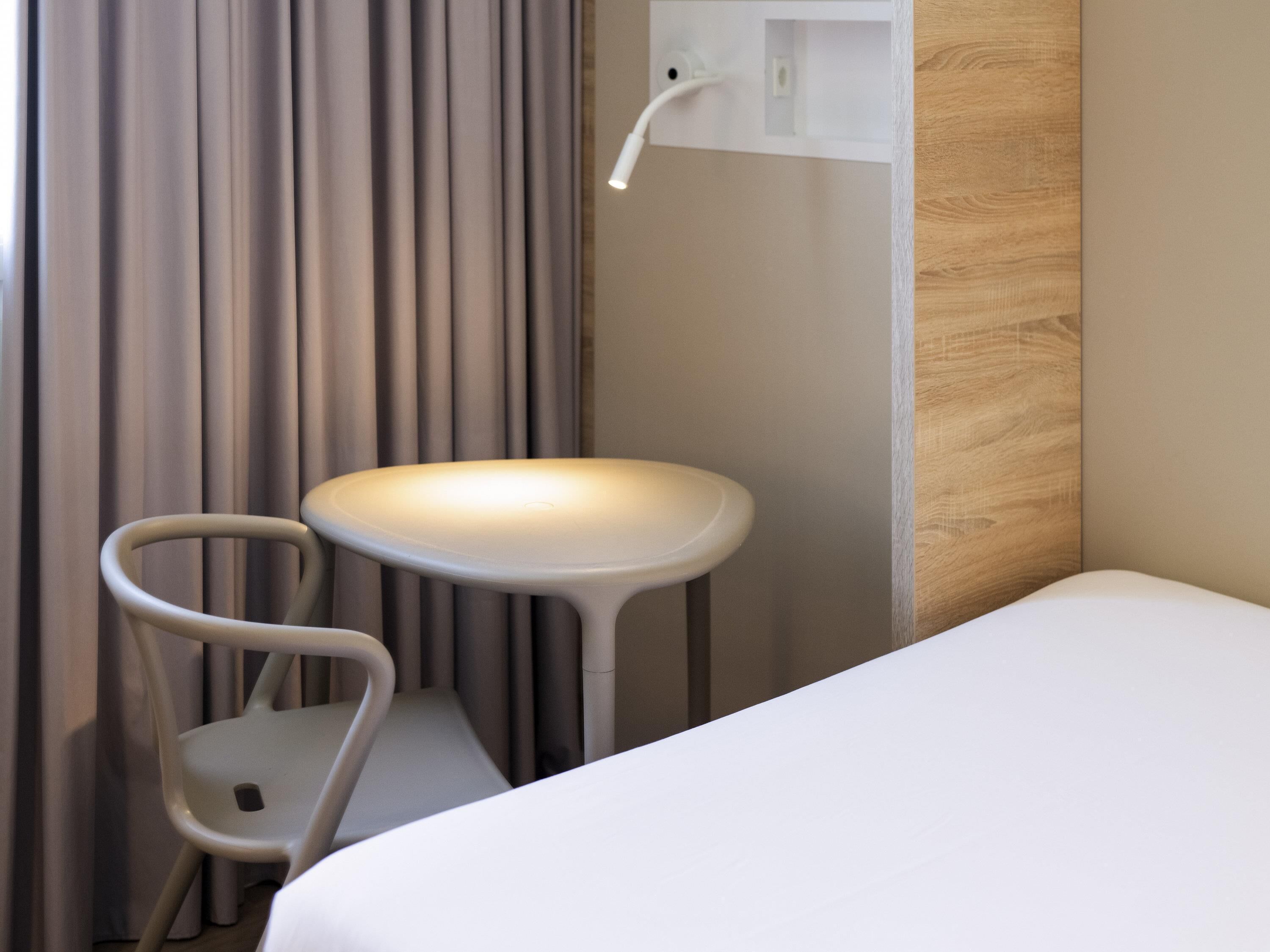 A modern hotel room with a bed, a chair, and a small white table with a light.