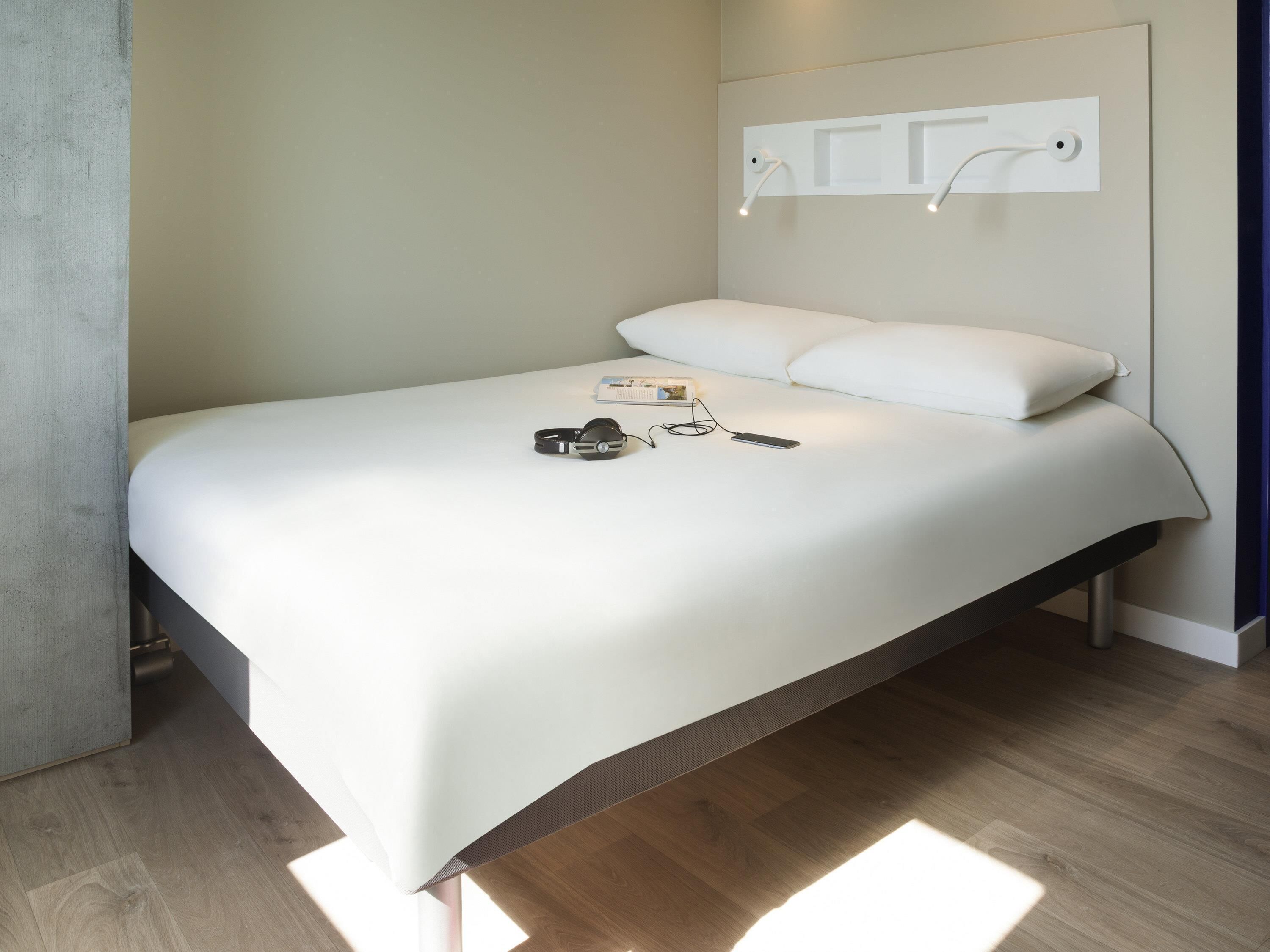 A modern hotel room with a bed, bedside table, and a wall-mounted light fixture.