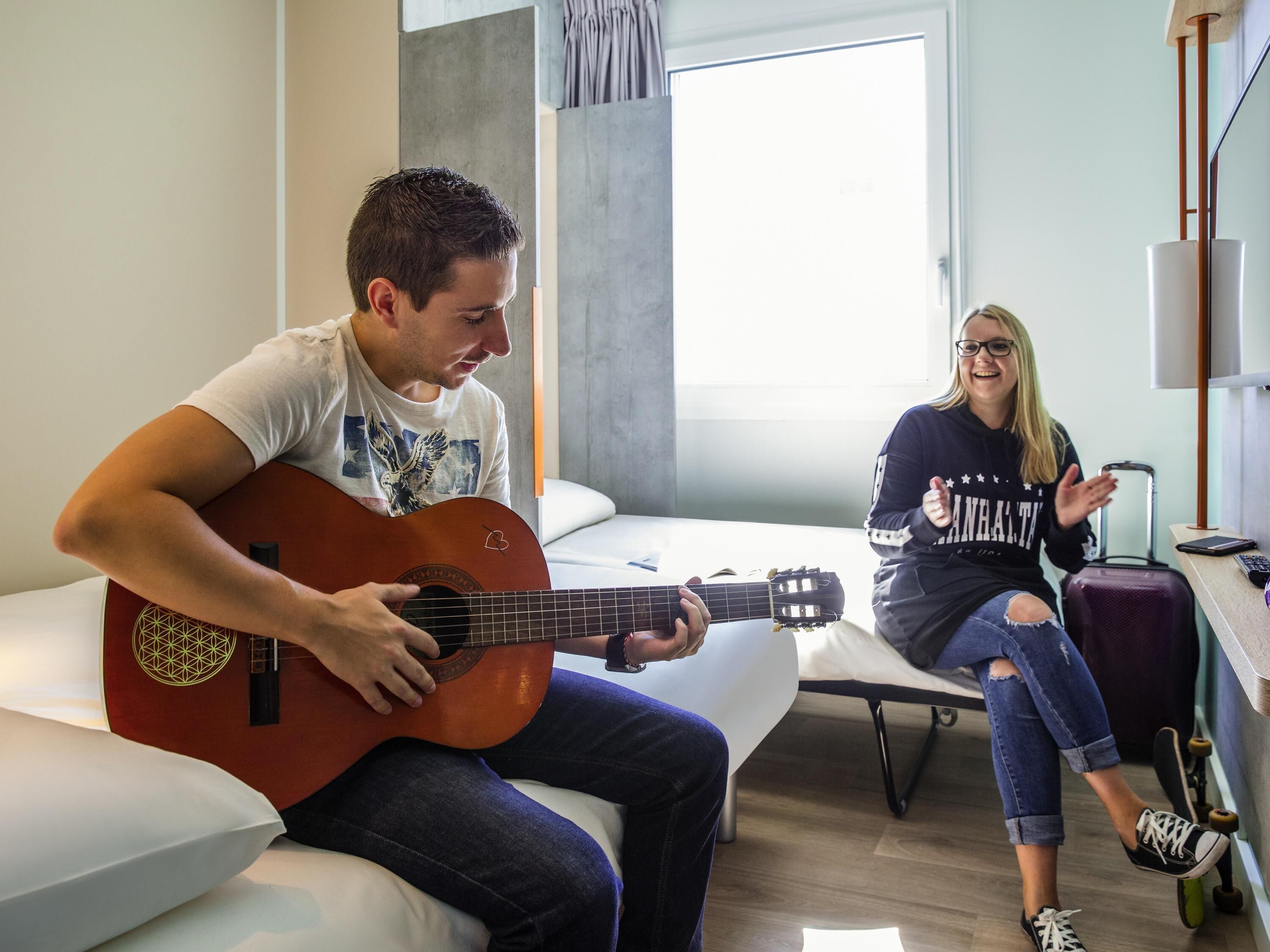 A man playing an acoustic guitar on a bed in a hotel room with a woman sitting on a couch, smiling and clapping.