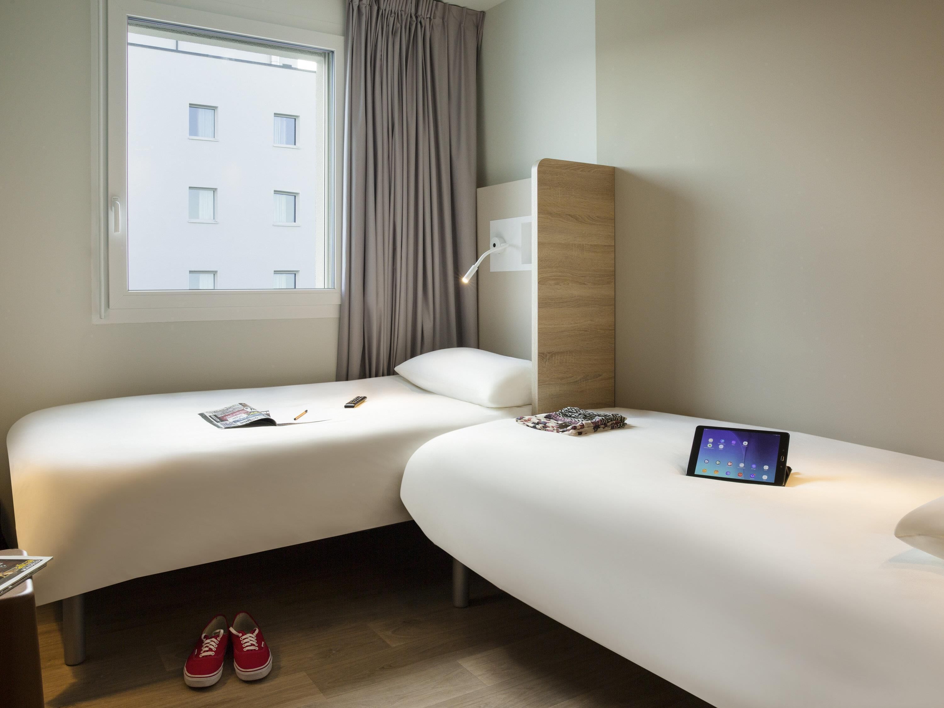 Quarto de hotel moderno com duas camas, uma janela com cortinas, cabeceira de madeira, mesa de cabeceira e um tablet sobre uma das camas.
