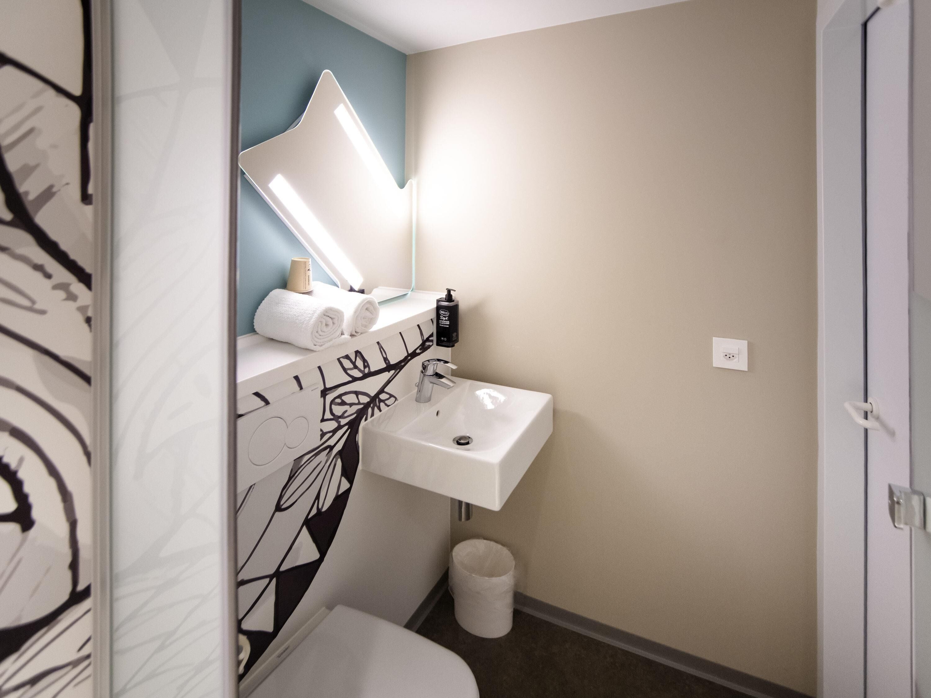 A small bathroom with a white sink, a mirror, and a toilet. There is a towel rack with towels and a bottle of soap on the sink.