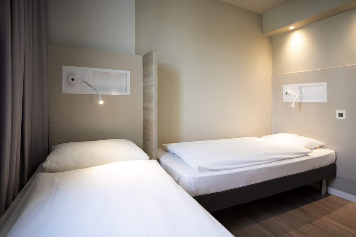 Une chambre d’hôtel avec deux lits simples, une tête de lit en bois, une lampe de lecture fixée au mur et une étagère intégrée.
