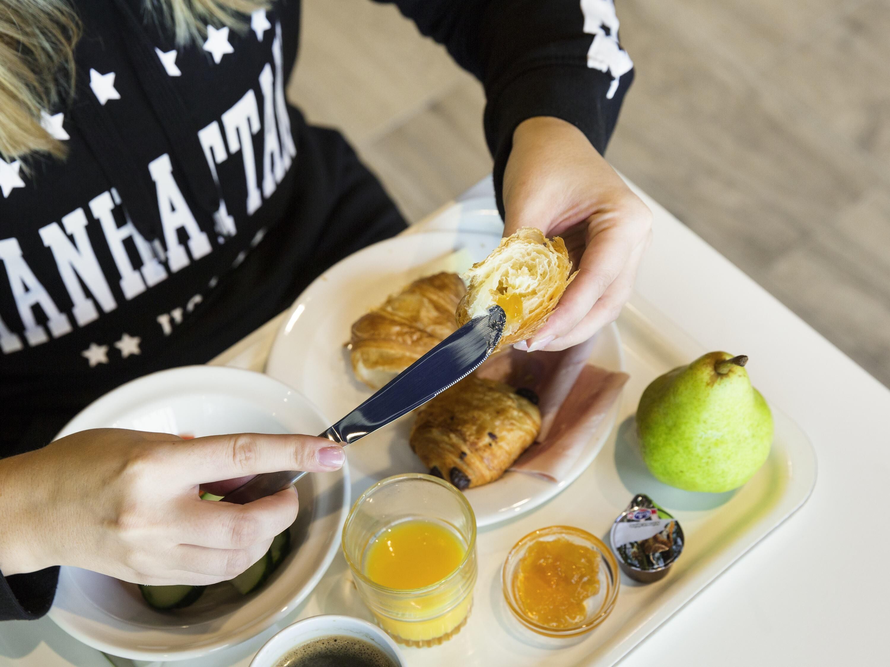 Une personne tartine du beurre sur une pâtisserie à l’aide d’un couteau, avec une poire et deux petits récipients contenant des condiments, le tout sur un plateau.