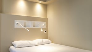 A single bed with white bedding, a wall-mounted light fixture, and a power outlet.