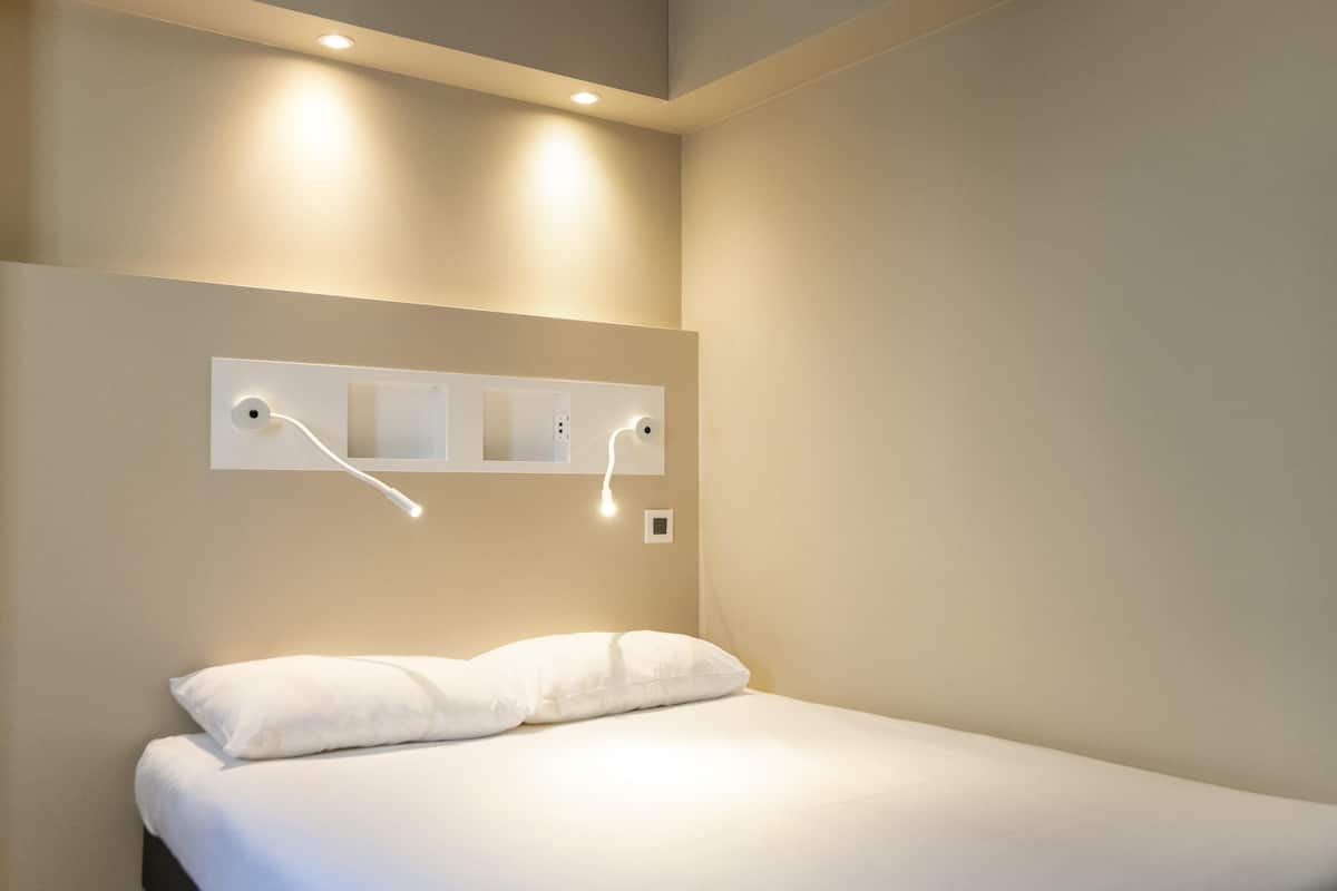 Un lit simple avec une literie blanche, un luminaire fixé au mur et une prise de courant.