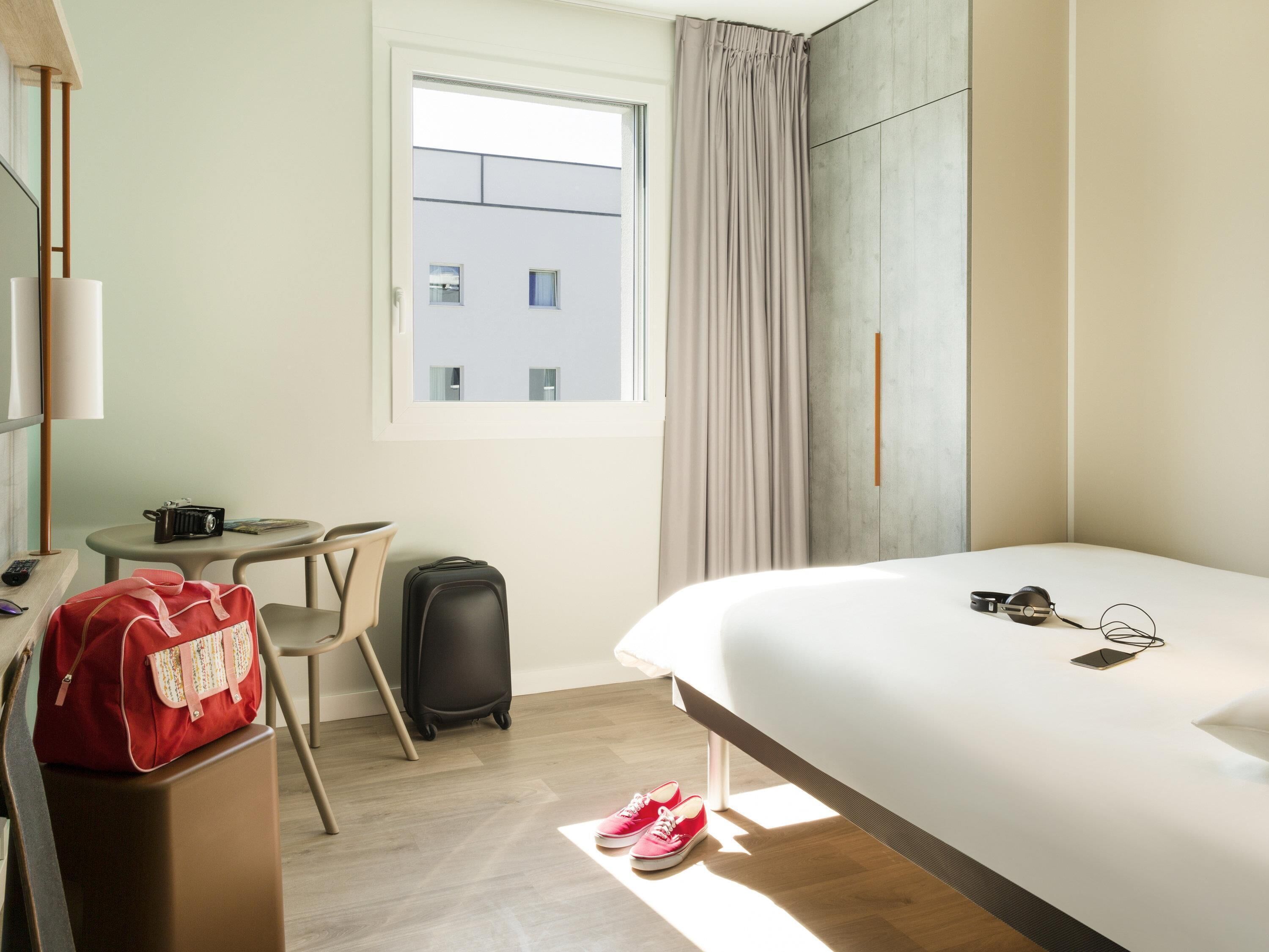Een hotelkamer met een bed, een bureau, een stoel, een koffer, een rode tas en een paar schoenen.