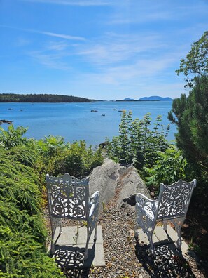 Two ornate metal chairs facing a body of water with boats, surrounded by greenery and a clear sky.