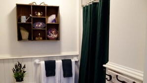 A bathroom with a wooden shelf holding decorative items, a white radiator, a green shower curtain, and two towels hanging on a towel rack.