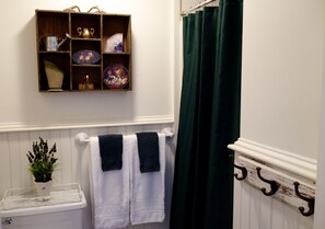 A bathroom with a wooden shelf holding decorative items, a white radiator, a green shower curtain, and two towels hanging on a towel rack.