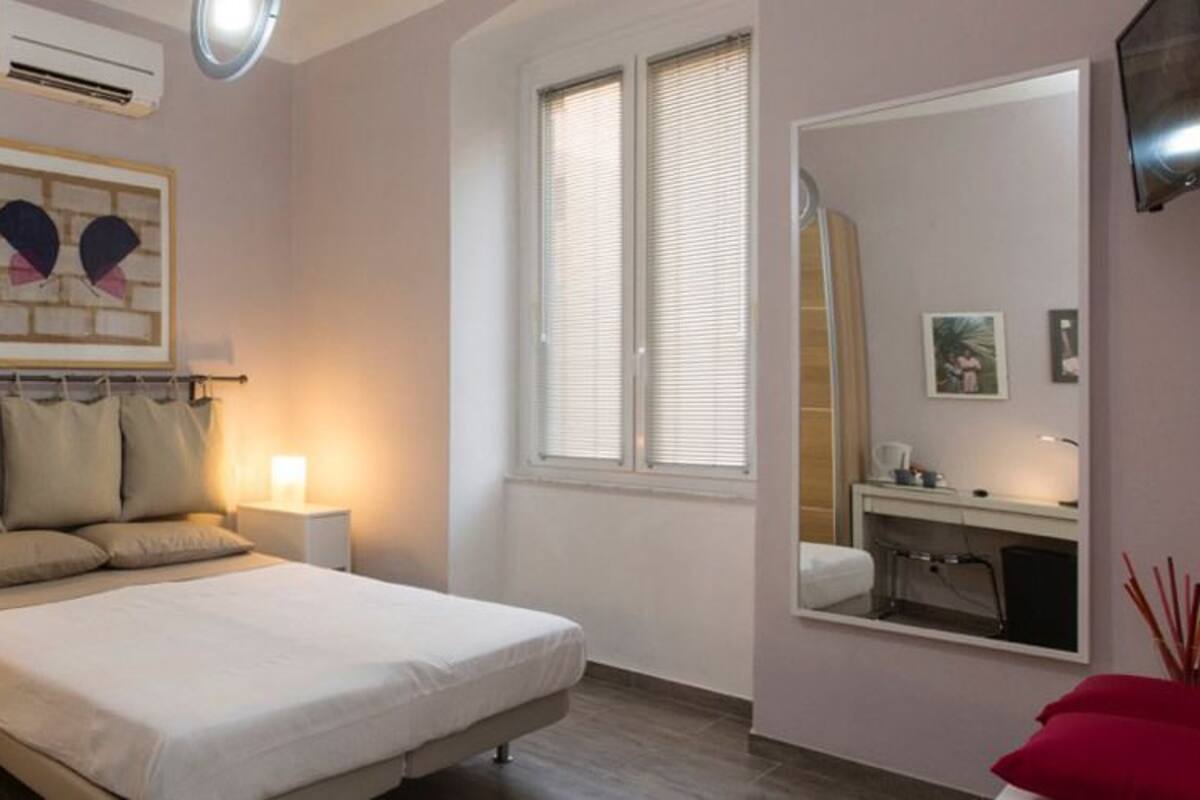 Un dormitorio con cama, mesitas de noche, una ventana, un espejo y televisor montado en la pared.