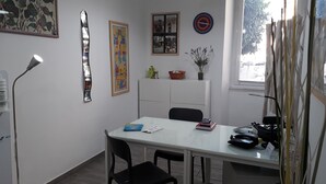 Une pièce avec une table blanche, des chaises noires, une lampe et divers éléments de décoration murale, notamment des tableaux encadrés et une horloge.