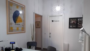 Un bureau moderne avec un bureau blanc, un ordinateur, un téléphone et divers fournitures de bureau. On peut voir un tableau abstrait encadré et une porte avec une pancarte.