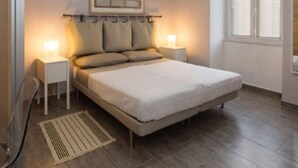 Une chambre à coucher comprenant un lit, des tables de chevet, un climatiseur fixé au mur, une fenêtre avec des stores et une porte.