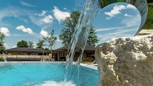 Een helderblauw zwembad met een waterval, omgeven door een houten terras en een huis op de achtergrond.