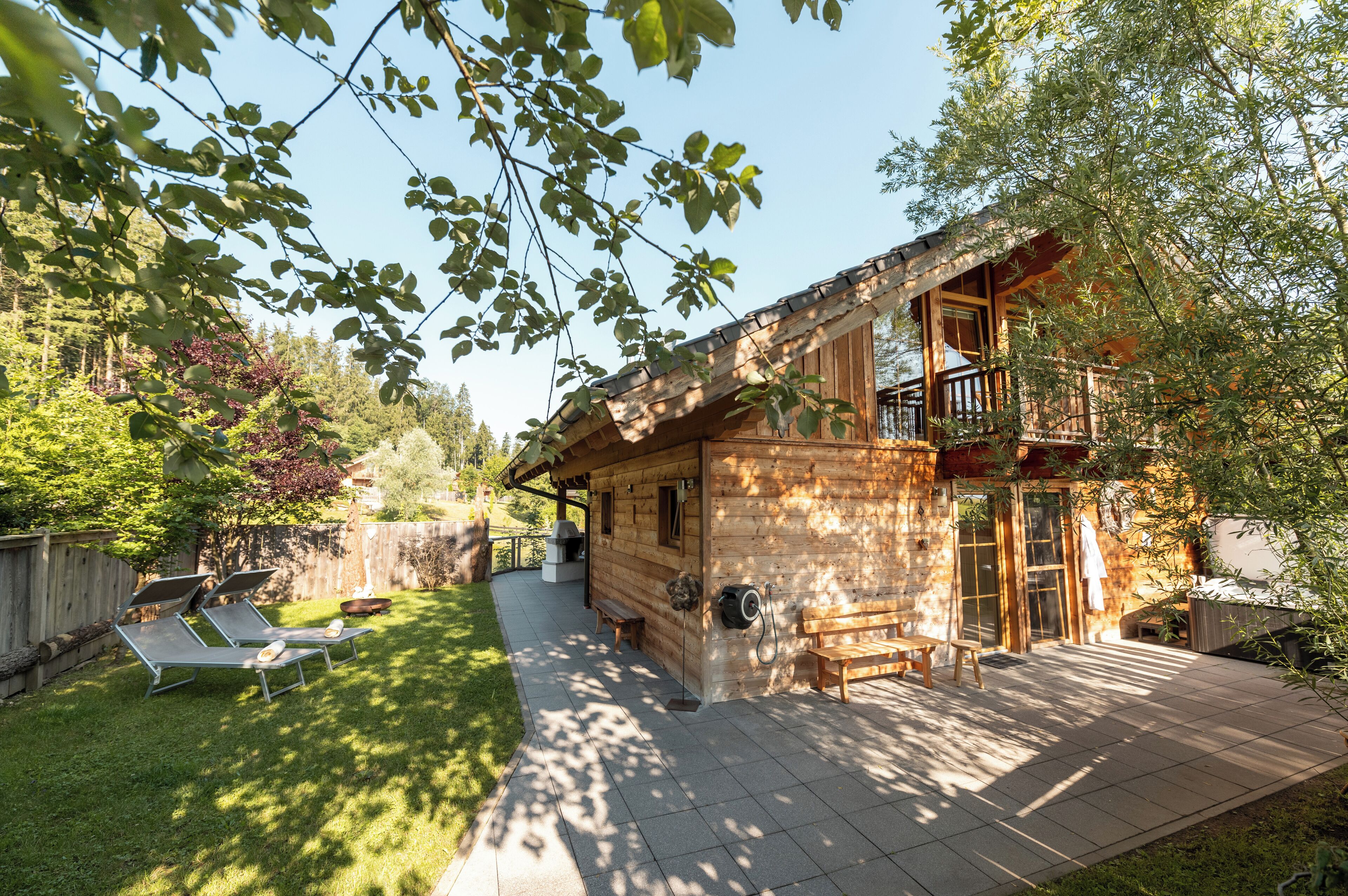 A wooden cabin with a balcony, a patio with a table and chairs, and a lawn area with sun loungers.