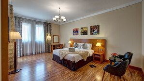 A hotel room with a large bed, two bedside tables with lamps, a desk with a chair, a small table with flowers, and a chandelier.