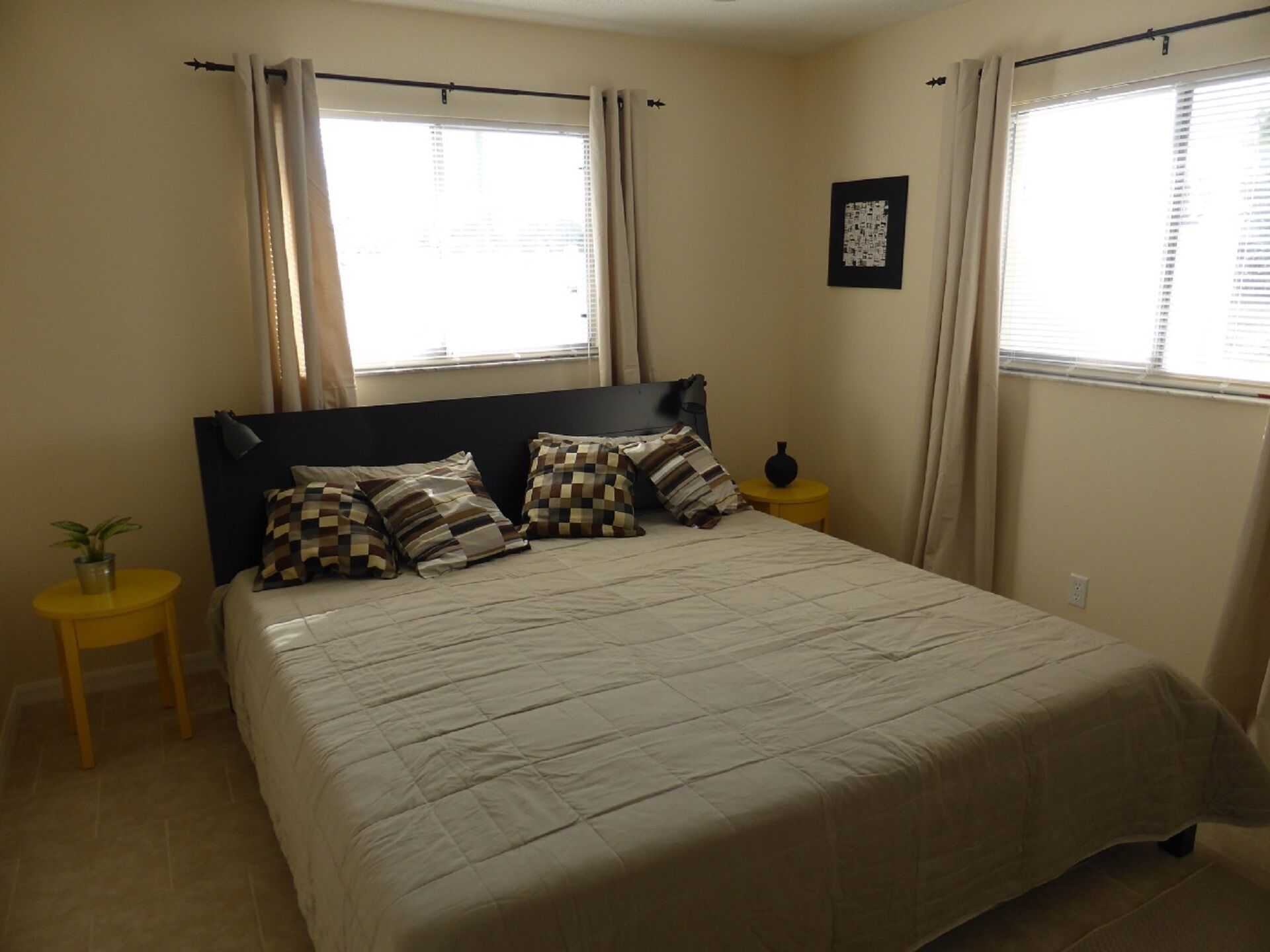 A bedroom with a large bed, two bedside tables, a potted plant, and curtains on the windows.