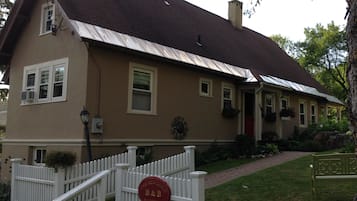 Welcome to The Red Door B&B, a charming two-story home surrounded by a lush lawn and trees, with a cozy porch and a white picket fence.