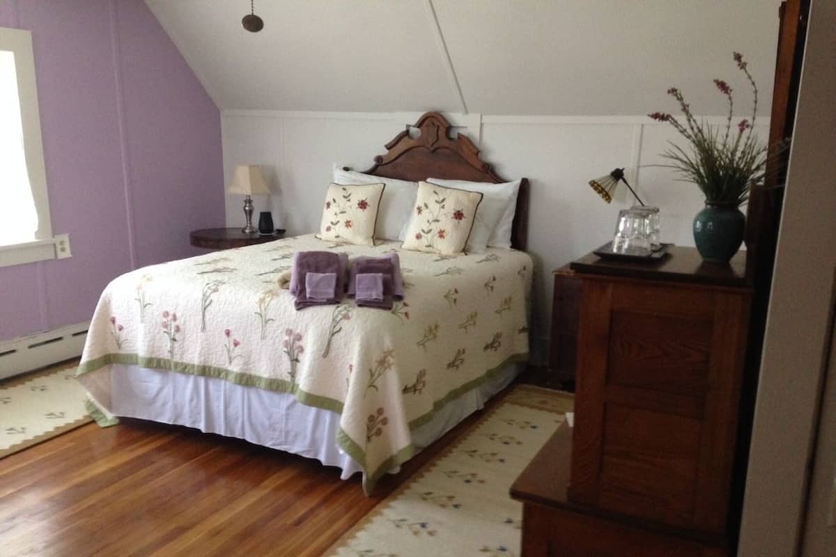 A bedroom with a wooden floor, a bed with floral bedding, a nightstand, a lamp, and a dresser with a vase of flowers.