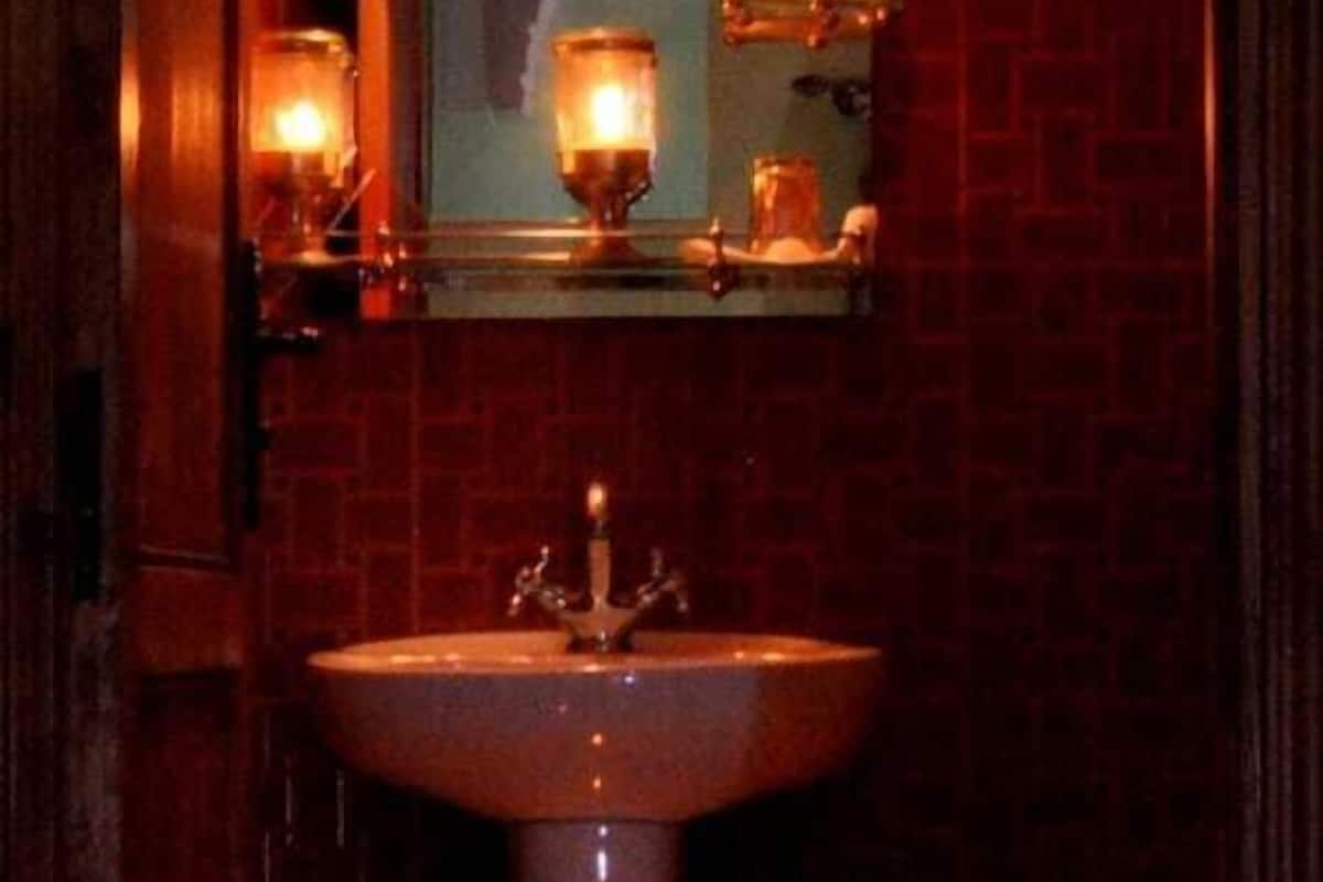 A narrow, dimly lit bathroom with a large sink, a mirror above it, and several candles providing light.
