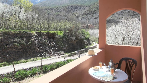 Un balcón con mesa lista para el desayuno, con vista a un paisaje montañoso con árboles y un muro de piedra.