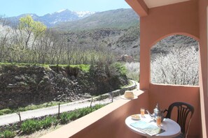 Un balcon avec une table dressée pour le petit-déjeuner, offrant une vue sur un paysage montagneux parsemé d’arbres et bordé d’un mur de pierres.