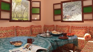 Un comedor con una mesa dispuesta para dos personas, con mantel estampado, un libro, una tetera, un plato de comida y una taza.