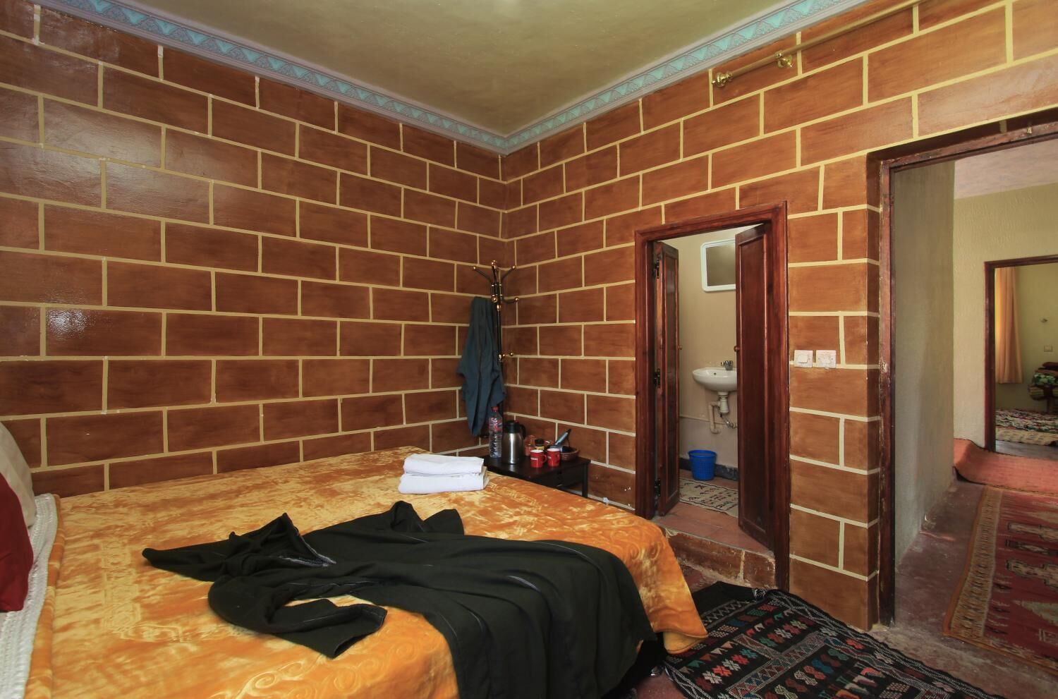 A room with a brick wall, a bed with a patterned bedspread, a suitcase, and a blue bucket.