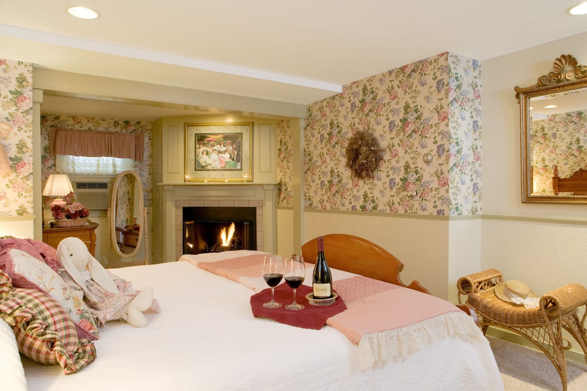 A bedroom with a fireplace, floral wallpaper, a bed with pillows, a wicker chair, and two wine glasses.