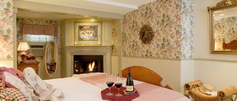 A bedroom with a fireplace, floral wallpaper, a bed with pillows, a wicker chair, and two wine glasses.