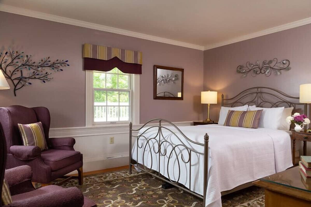 A bedroom with a metal bed frame, a purple armchair, a window with a striped valance, and a decorative wall piece.