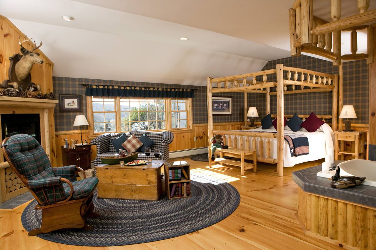 A log cabin-style room with a large bed, a rocking chair, a fireplace, and a wooden ceiling.