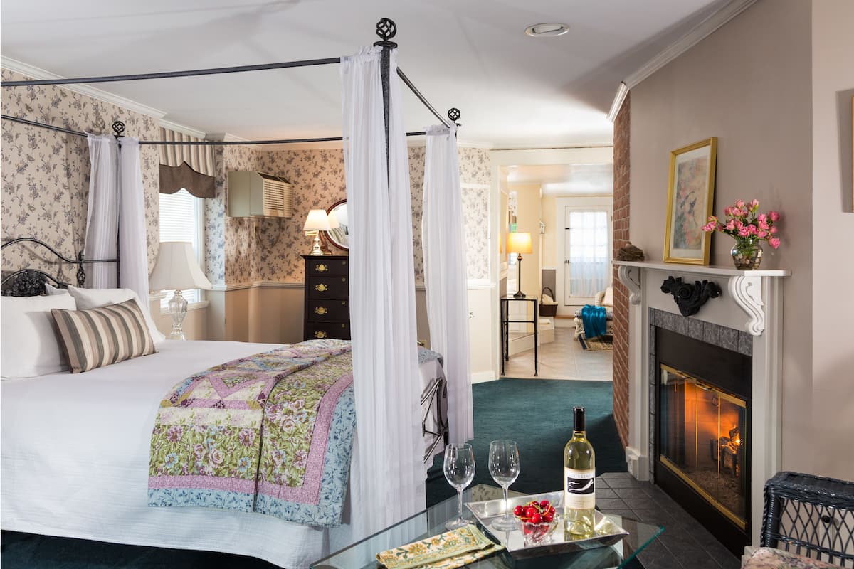 A four-poster bed with a canopy, a fireplace, and a glass table with wine glasses and a bottle.