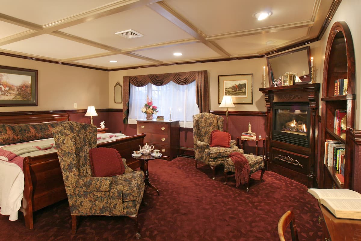 A room with a bed, two armchairs, a small table with a lamp, a fireplace, and a bookshelf.