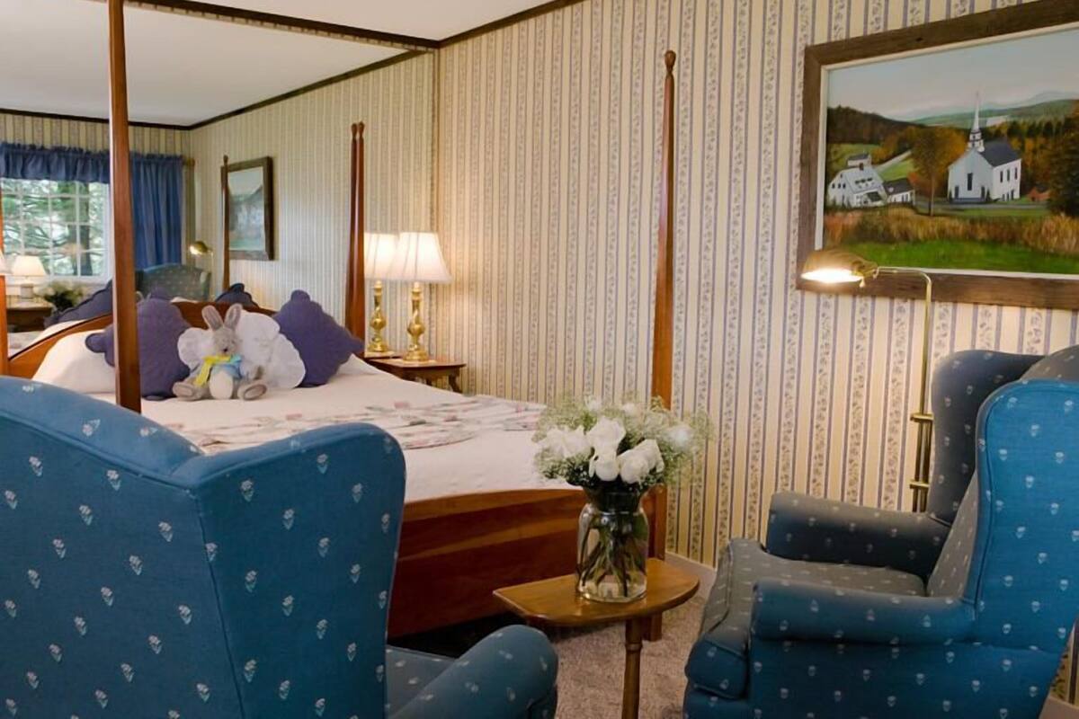 A hotel room with a four-poster bed, a large painting of a countryside scene, and a vase of white flowers on a side table.