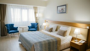 A hotel room with a large bed, two blue armchairs, a small round table, and a nightstand with a lamp and phone.