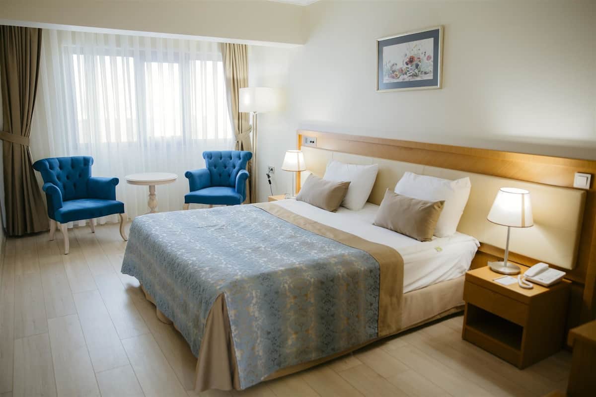 Habitación de hotel con una cama grande, dos sillones azules, una mesita redonda y una mesita de noche con lámpara y teléfono.