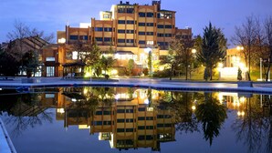 A multi-story hotel with a reflection in a calm water body, surrounded by trees and a well-lit street.