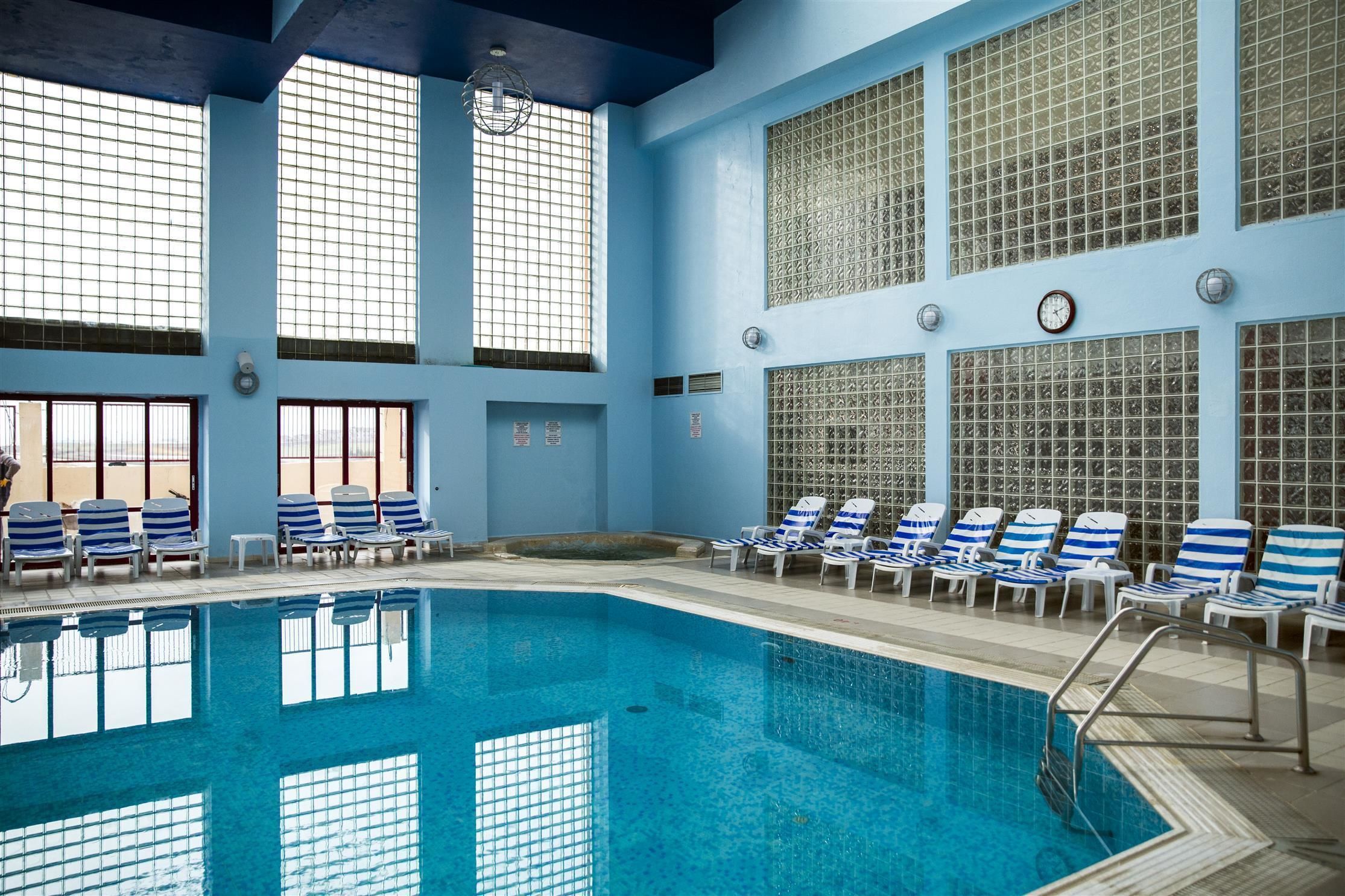 An indoor swimming pool with blue-tiled walls, large windows, and sunlit areas with lounge chairs.