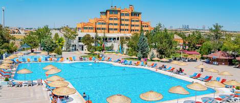 A large swimming pool surrounded by lounge chairs and umbrellas, with a hotel building in the background.