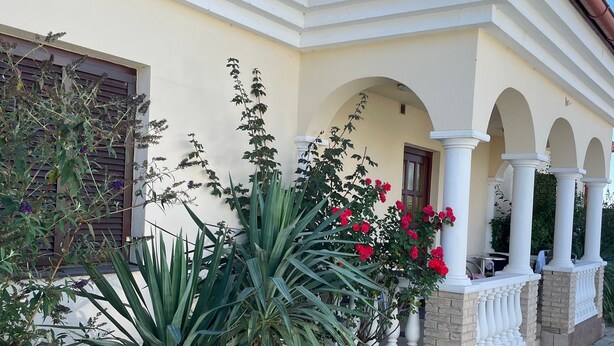 A building with a tiled roof, a covered porch with columns, and a garden with various plants and flowers.