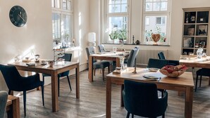 Un comedor bien iluminado con mesas y sillas de madera, ventanas amplias y una estantería con diversos objetos.