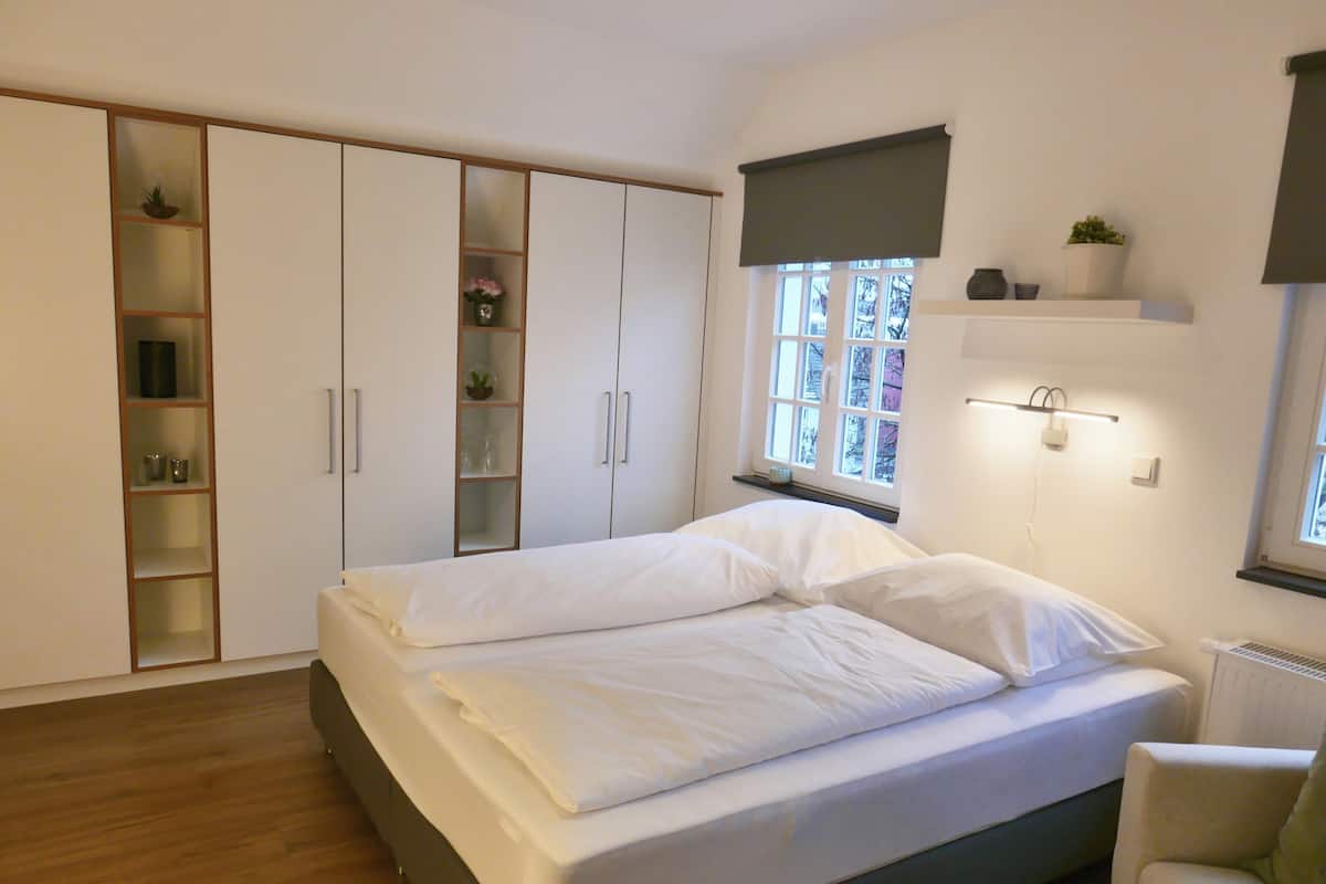 A bedroom with a large bed, a wardrobe, a shelf with plants, and a window with a view.