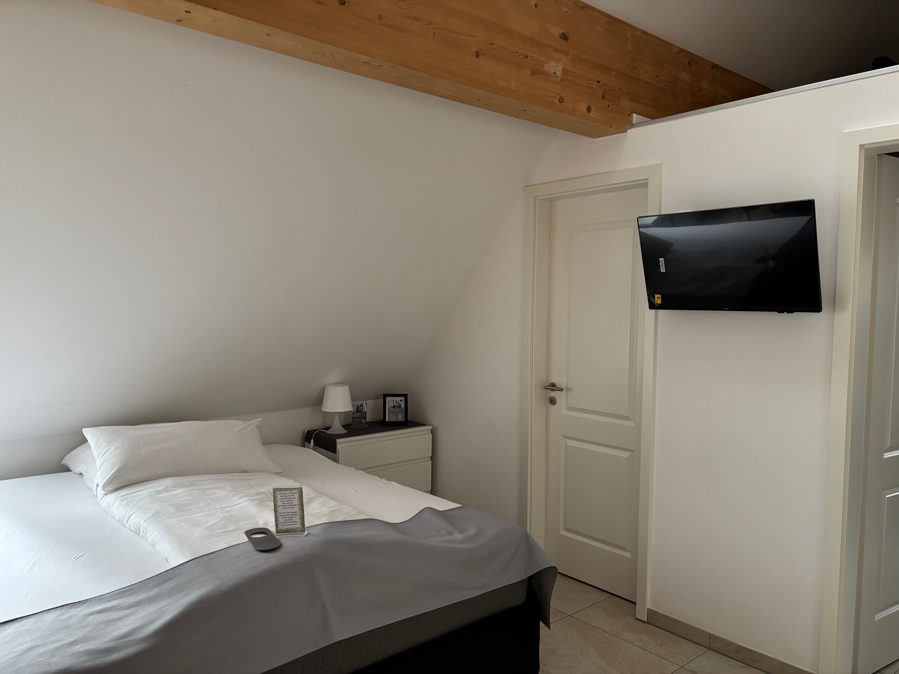 A bedroom with a bed, a bedside table with a lamp, a TV mounted on the wall, and a door.