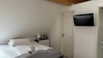 A bedroom with a bed, a bedside table with a lamp, a TV mounted on the wall, and a door.