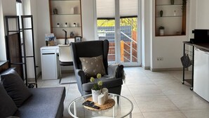 Una sala moderna con sofá, sillón y mesa de centro. Hay estantes con objetos decorativos y una vista al exterior a través de una puerta de vidrio.
