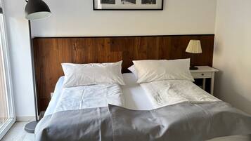 Ein ordentlich bezogenes Doppelbett mit weißer und grauer Bettwäsche, ein Holz-Kopfteil, ein gerahmtes Bild über dem Bett und eine Nachttischlampe auf einem Nachttisch.