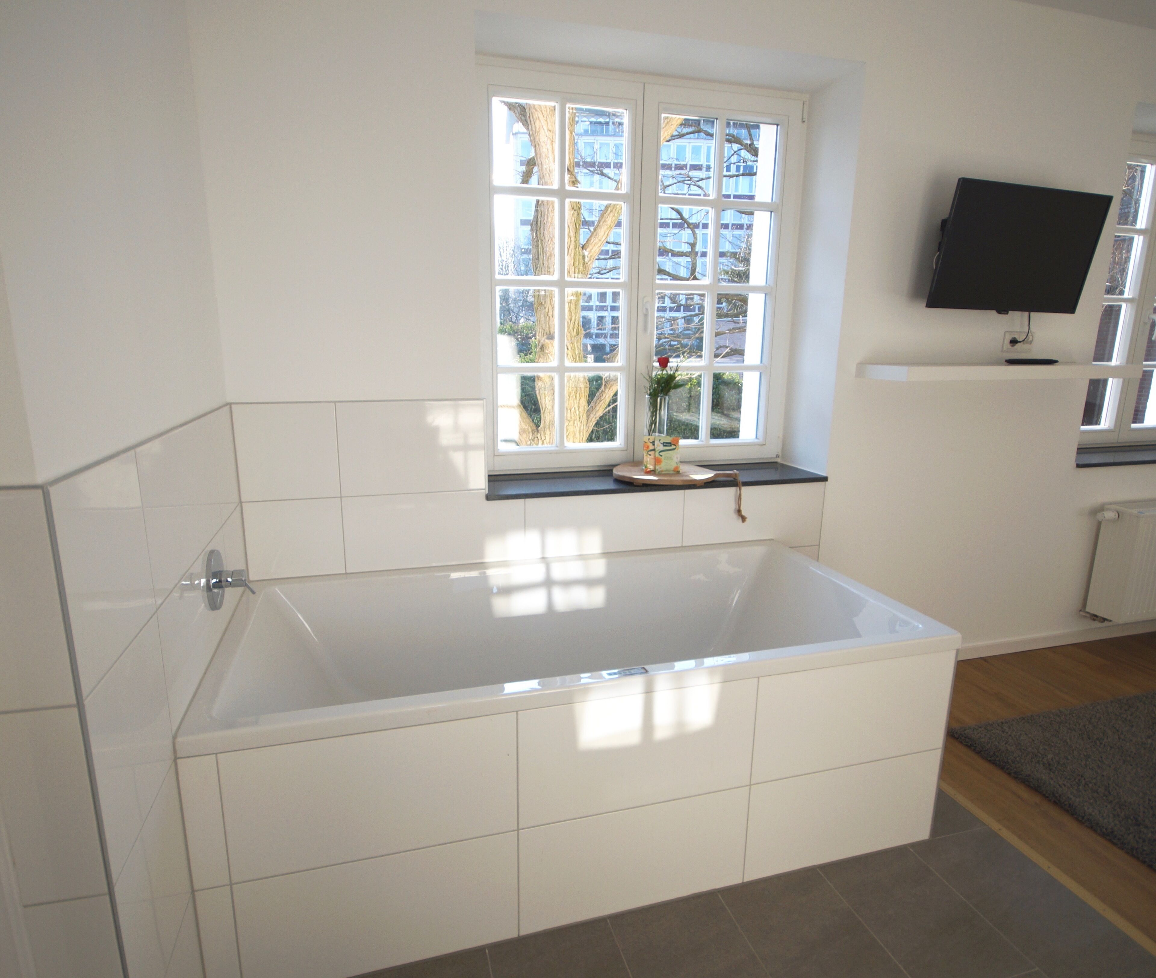 Ein modernes Badezimmer mit einer großen Badewanne, einem Fenster mit Blick auf Gebäude und einem an der Wand befestigten Flachbildfernseher.